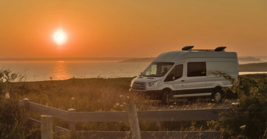 Ford Transit camper van parked near a scenic coastline at sunset, with the sun casting a warm glow over the landscape.